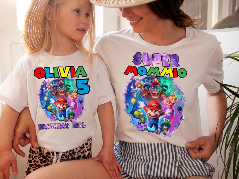 May include: White t-shirts with colorful Mario-themed graphics. One shirt says "Olivia 5 Birthday Girl" and the other says "Super Mommio". The graphics feature Mario, Luigi, and other characters.