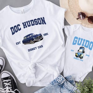 May include: Two white t-shirts with blue text and images. The larger shirt says "Doc Hudson Est 1951 Disney Car" with a blue car image. The smaller shirt says "Guido Est 2008 Disney Car" with a blue car image.