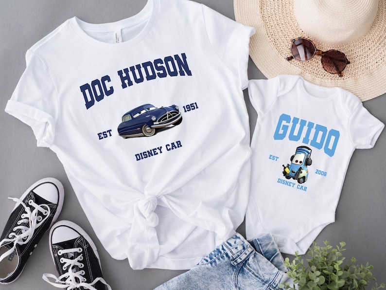 May include: Two white t-shirts with blue text and images. The larger shirt says "Doc Hudson Est 1951 Disney Car" with a blue car image. The smaller shirt says "Guido Est 2008 Disney Car" with a blue car image.