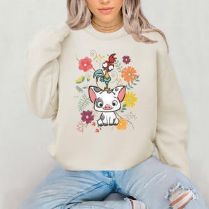 May include: A cream-coloured sweatshirt featuring a cartoon image of a pig and a rooster surrounded by flowers. The pig is white with black spots and the rooster is red, yellow, and green.