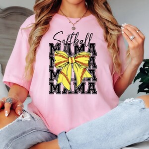 K&ouml;nnte beinhalten: Pinkes T-Shirt mit dem Text "Softball Mama" in schwarzen Buchstaben und einer gelben und roten Softball-Schleife.