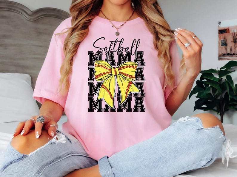K&ouml;nnte beinhalten: Pinkes T-Shirt mit dem Text "Softball Mama" in schwarzen Buchstaben und einer gelben und roten Softball-Schleife.