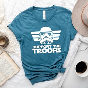 May include: A teal blue t-shirt featuring a white graphic of a Star Wars Stormtrooper helmet with wings and the text "Support the Troops".