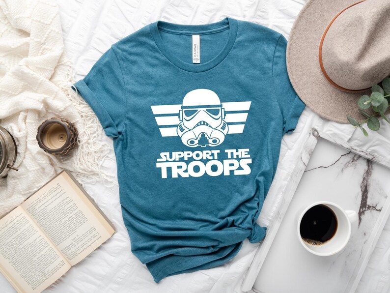 May include: A teal blue t-shirt featuring a white graphic of a Star Wars Stormtrooper helmet with wings and the text "Support the Troops".