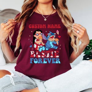 Pu&ograve; includere: T-shirt bordeaux con grafica di Lilo e Stitch che mangiano gelati. Il testo "CUSTOM NAME" &egrave; sopra l'immagine e "BESTIE FOREVER" sotto. La maglietta ha girocollo e maniche corte.