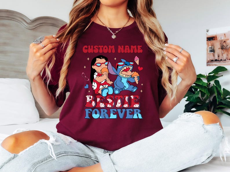 Pu&ograve; includere: T-shirt bordeaux con grafica di Lilo e Stitch che mangiano gelati. Il testo "CUSTOM NAME" &egrave; sopra l'immagine e "BESTIE FOREVER" sotto. La maglietta ha girocollo e maniche corte.