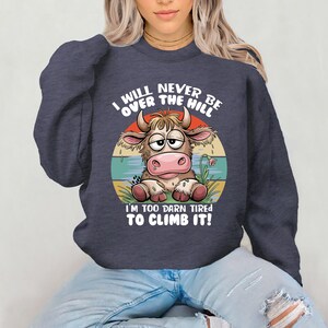 Puede incluir: Una sudadera azul marino con una vaca de dibujos animados y el texto "I will never be over the hill I'm too darn tired to climb it!"