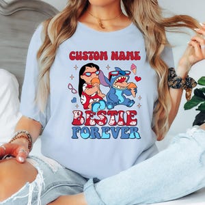Pu&ograve; includere: T-shirt azzurra con una grafica di due personaggi dei cartoni animati, uno rosso e uno blu, che mangiano gelati. Il testo "CUSTOM NAME" &egrave; sopra i personaggi e "BESTIE FOREVER" sotto. La maglietta &egrave; di colore azzurro.