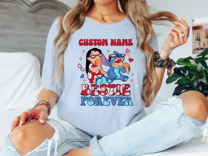 Pu&ograve; includere: T-shirt azzurra con una grafica di due personaggi dei cartoni animati, uno rosso e uno blu, che mangiano gelati. Il testo "CUSTOM NAME" &egrave; sopra i personaggi e "BESTIE FOREVER" sotto. La maglietta &egrave; di colore azzurro.