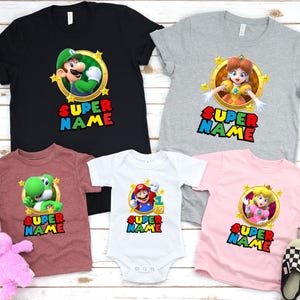 May include: Set of five t-shirts featuring characters from the Super Mario video game franchise. The shirts are in various colors, including black, gray, pink, white, and maroon. Each shirt features a different character, including Mario, Luigi, Peach, Yoshi, and Daisy. The shirts all have the text "SUPER NAME" printed on them.