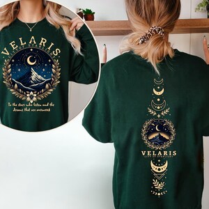 May include: Green long-sleeved sweatshirt with a gold graphic print of a mountain range and crescent moon with the text "VELARIS CITY OF STARLIGHT" and "To the stars who listen and the dreams that are answered". The back of the sweatshirt has a gold graphic print of a mountain range, crescent moon, and stars with the text "VELARIS CITY OF STARLIGHT".