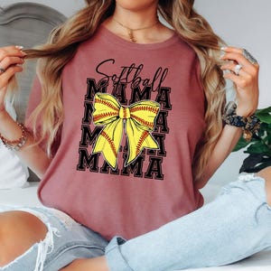 K&ouml;nnte beinhalten: Ein maronfarbenes T-Shirt mit dem Aufdruck "Softball Mama". Der Text ist von einer gelben und roten Softball-Schleife umgeben.