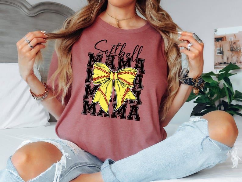 K&ouml;nnte beinhalten: Ein maronfarbenes T-Shirt mit dem Aufdruck "Softball Mama". Der Text ist von einer gelben und roten Softball-Schleife umgeben.