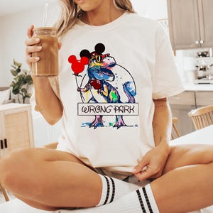 May include: White t-shirt with a colorful graphic of a dinosaur wearing Mickey Mouse ears and sunglasses holding a red balloon. The text "Wrong Park" is printed below the dinosaur.