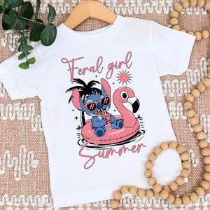 以下が含まれることがあります： 白いTシャツ。サングラスをかけた漫画のキャラクターがピンクのフラミンゴフロートに乗っています。「Feral girl summer」という文字がピンク色で書かれています。Tシャツは木製のハンガーにかかっています。