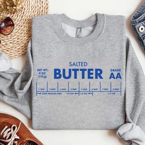 May include: A heather gray sweatshirt with the words "SALTED BUTTER" in blue, along with a measurement guide. The sweatshirt also features the text "NET WT. 4 OZ (113g)" and "GRADE AA".