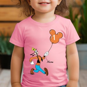 May include: Pink t-shirt with a cartoon image of Goofy holding a Mickey Mouse balloon with the number 1 on it. The text "Name" is below the image.