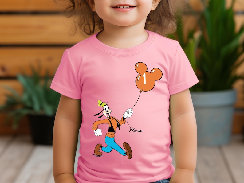 May include: Pink t-shirt with a cartoon image of Goofy holding a Mickey Mouse balloon with the number 1 on it. The text "Name" is below the image.