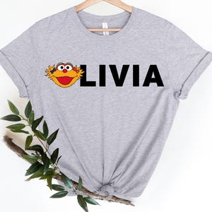 May include: A light gray t-shirt featuring a cartoon character's face and the name "LIVIA" in bold, black letters. The character has yellow fur, a red mouth, and large, round eyes. The shirt is made of a soft material.