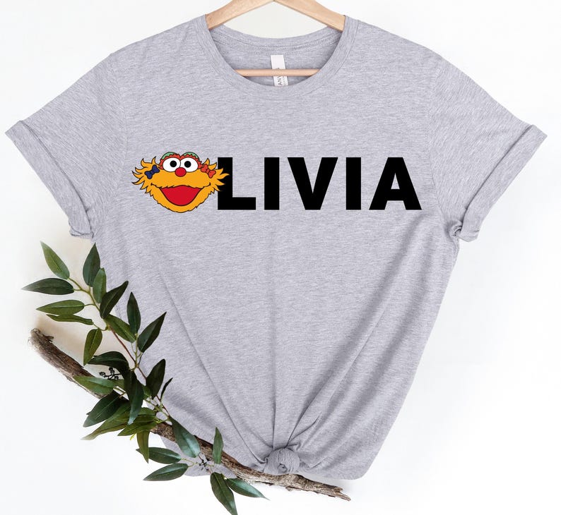 May include: A light gray t-shirt featuring a cartoon character's face and the name "LIVIA" in bold, black letters. The character has yellow fur, a red mouth, and large, round eyes. The shirt is made of a soft material.