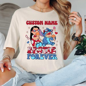 Pu&ograve; includere: T-shirt color crema con una grafica di due personaggi dei cartoni animati, uno rosso e uno blu, che tengono coni gelato. Il testo "CUSTOM NAME" e "BESTIE FOREVER" sono sopra e sotto i personaggi.
