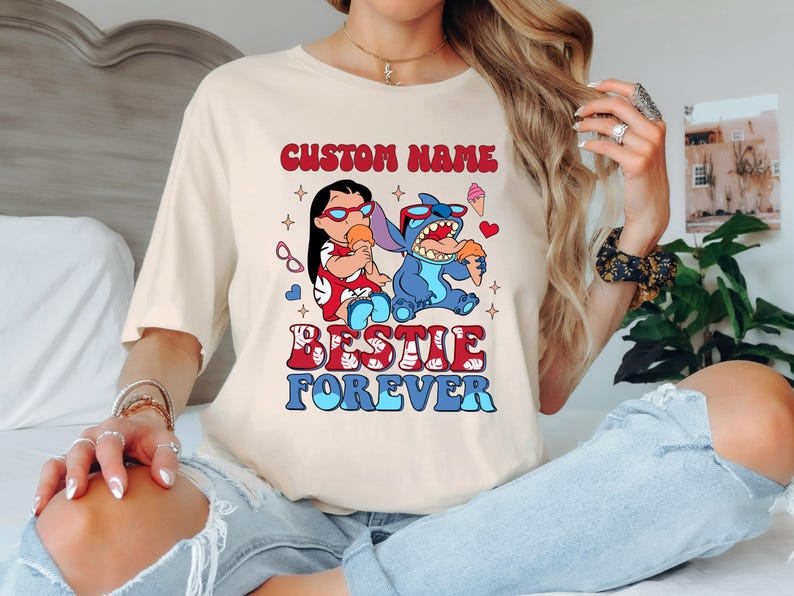 Pu&ograve; includere: T-shirt color crema con una grafica di due personaggi dei cartoni animati, uno rosso e uno blu, che tengono coni gelato. Il testo "CUSTOM NAME" e "BESTIE FOREVER" sono sopra e sotto i personaggi.