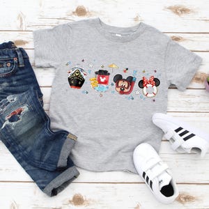 May include: A grey t-shirt featuring a colourful graphic of Mickey Mouse, Minnie Mouse and Donald Duck characters on a boat. The characters are wearing swimsuits and holding beach accessories. The graphic is surrounded by stars and other nautical elements.