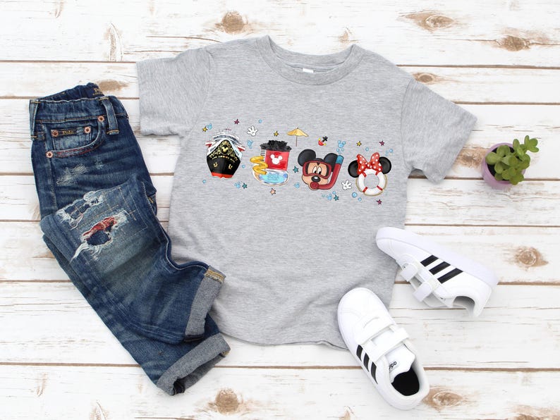 May include: A grey t-shirt featuring a colourful graphic of Mickey Mouse, Minnie Mouse and Donald Duck characters on a boat. The characters are wearing swimsuits and holding beach accessories. The graphic is surrounded by stars and other nautical elements.