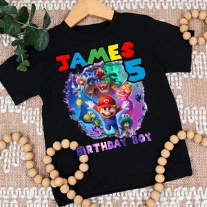 May include: Black t-shirt featuring a colorful graphic of video game characters, including Mario, Bowser, and Princess Peach. The shirt has the text "JAMES 5 BIRTHDAY BOY" in vibrant colors. The shirt is displayed on a wooden hanger.