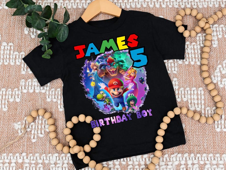 May include: Black t-shirt featuring a colorful graphic of video game characters, including Mario, Bowser, and Princess Peach. The shirt has the text "JAMES 5 BIRTHDAY BOY" in vibrant colors. The shirt is displayed on a wooden hanger.