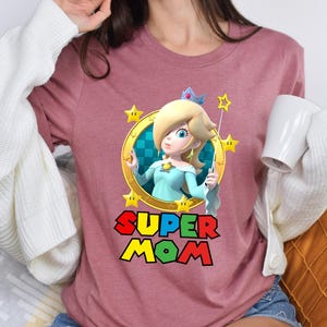 May include: A pink t-shirt with a graphic of Princess Peach from the Super Mario Bros. video game series. The graphic features Peach holding a magic wand and surrounded by stars. The text "SUPER MOM" is printed below the graphic.