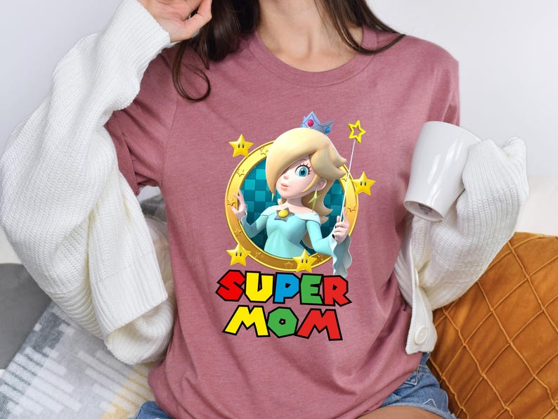 May include: A pink t-shirt with a graphic of Princess Peach from the Super Mario Bros. video game series. The graphic features Peach holding a magic wand and surrounded by stars. The text "SUPER MOM" is printed below the graphic.
