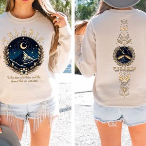 May include: A cream-coloured sweatshirt with a graphic design featuring a mountain range, crescent moon, and stars. The text "VELARIS CITY OF STARLIGHT" is printed in a gold-coloured font. The text "To the dust who gather, and the dreams that are awakened" is printed below the graphic.