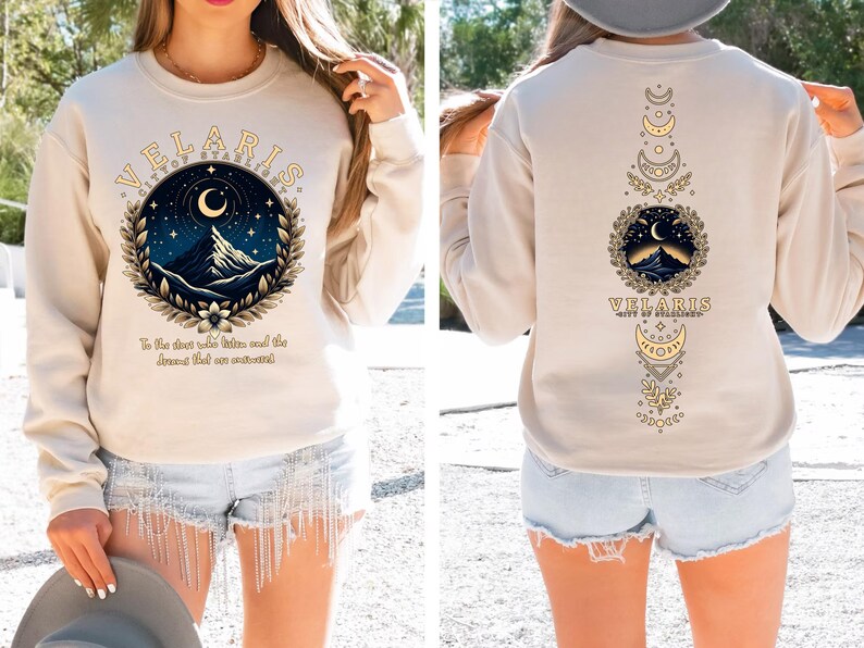 May include: A cream-coloured sweatshirt with a graphic design featuring a mountain range, crescent moon, and stars. The text "VELARIS CITY OF STARLIGHT" is printed in a gold-coloured font. The text "To the dust who gather, and the dreams that are awakened" is printed below the graphic.