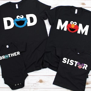 May include: Black family matching t-shirts and a onesie. The shirts say "DAD", "MOM", "BROTHER", and "SISTER" in white block letters. Each shirt has a Sesame Street character graphic: Cookie Monster, Elmo, and Abby Cadabby.