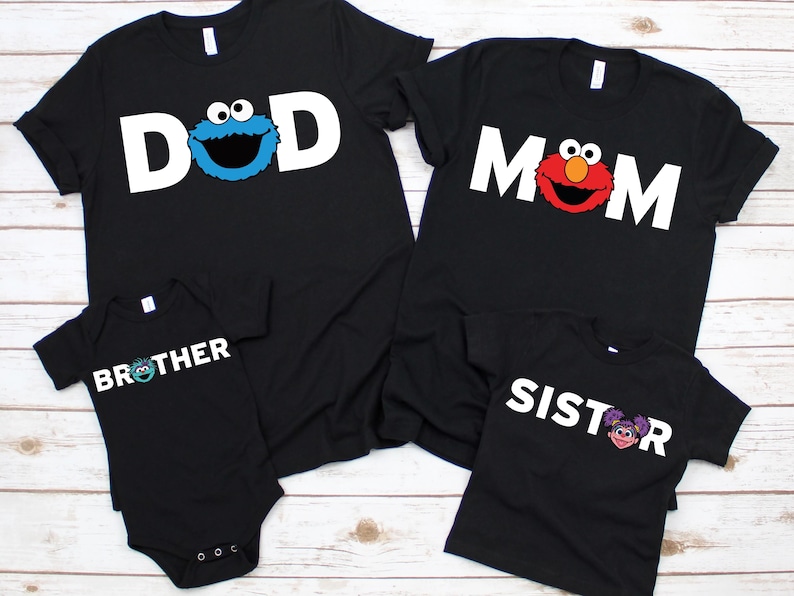May include: Black family matching t-shirts and a onesie. The shirts say "DAD", "MOM", "BROTHER", and "SISTER" in white block letters. Each shirt has a Sesame Street character graphic: Cookie Monster, Elmo, and Abby Cadabby.