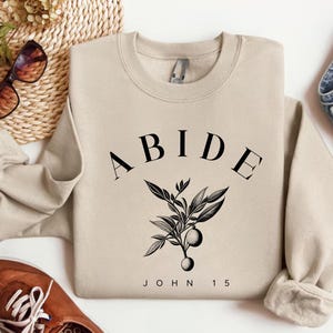 May include: A beige sweatshirt with the word "ABIDE" in black arched text above a black and white botanical illustration. The text "JOHN 15" is below the illustration. Sunglasses, shoes, and denim jeans are also in the image.