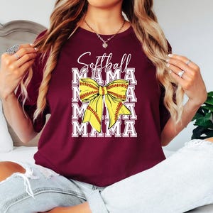 K&ouml;nnte beinhalten: Ein maroonfarbenes T-Shirt mit dem Text "Softball Mama", der viermal in Wei&szlig; wiederholt wird. Ein gelber Softball mit roten N&auml;hten ist in der Mitte des Shirts zu einem Bogen gebunden.