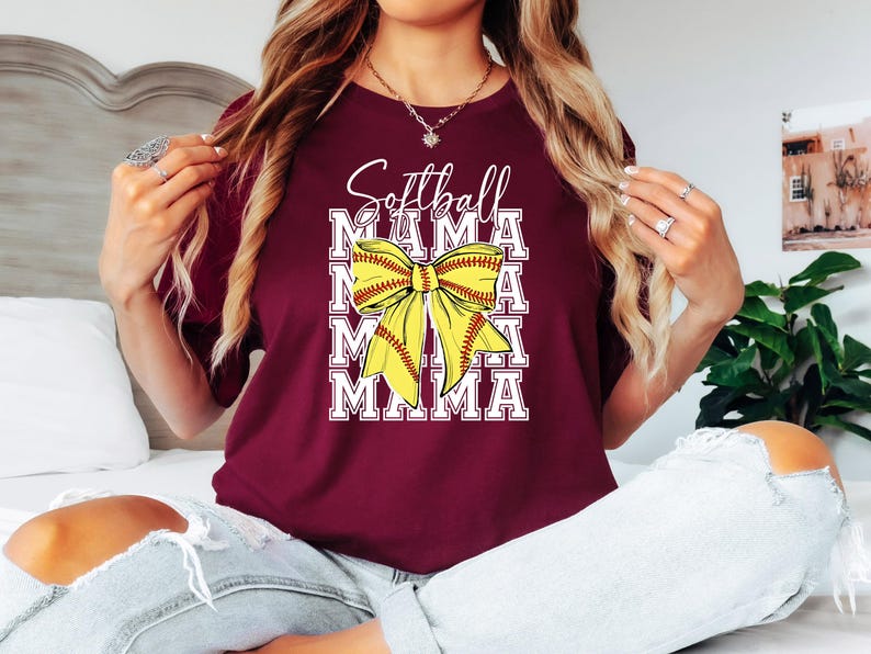 K&ouml;nnte beinhalten: Ein maroonfarbenes T-Shirt mit dem Text "Softball Mama", der viermal in Wei&szlig; wiederholt wird. Ein gelber Softball mit roten N&auml;hten ist in der Mitte des Shirts zu einem Bogen gebunden.
