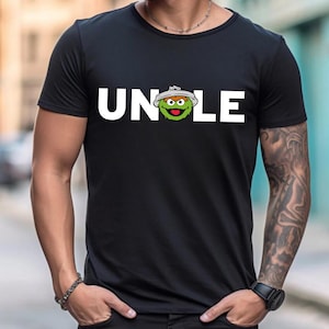 May include: Black t-shirt with the word "UNCLE" in white block letters. The "O" in "UNCLE" is replaced with a cartoon character's face. The character is green with a white hat.