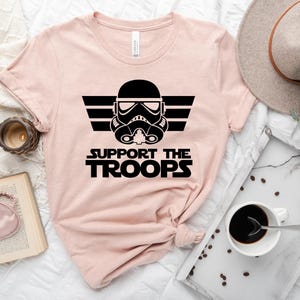 May include: A pink t-shirt featuring a black graphic of a Star Wars Stormtrooper helmet with wings and the text "SUPPORT THE TROOPS".