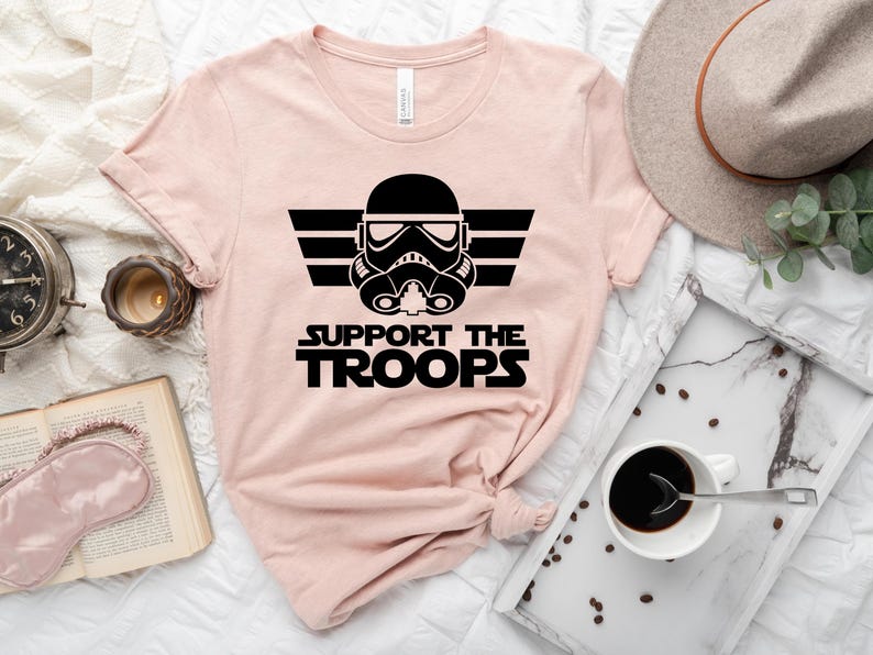 May include: A pink t-shirt featuring a black graphic of a Star Wars Stormtrooper helmet with wings and the text "SUPPORT THE TROOPS".