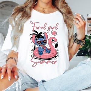 以下が含まれることがあります： 白いTシャツ。サングラスをかけた漫画のキャラクターが、ピンクのフラミンゴのフロートに乗っています。シャツにはピンクのスクリプトで「Feral girl summer」と書かれています。デザインにはヤシの木と太陽が含まれています。