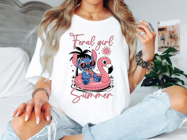 以下が含まれることがあります： 白いTシャツ。サングラスをかけた漫画のキャラクターが、ピンクのフラミンゴのフロートに乗っています。シャツにはピンクのスクリプトで「Feral girl summer」と書かれています。デザインにはヤシの木と太陽が含まれています。
