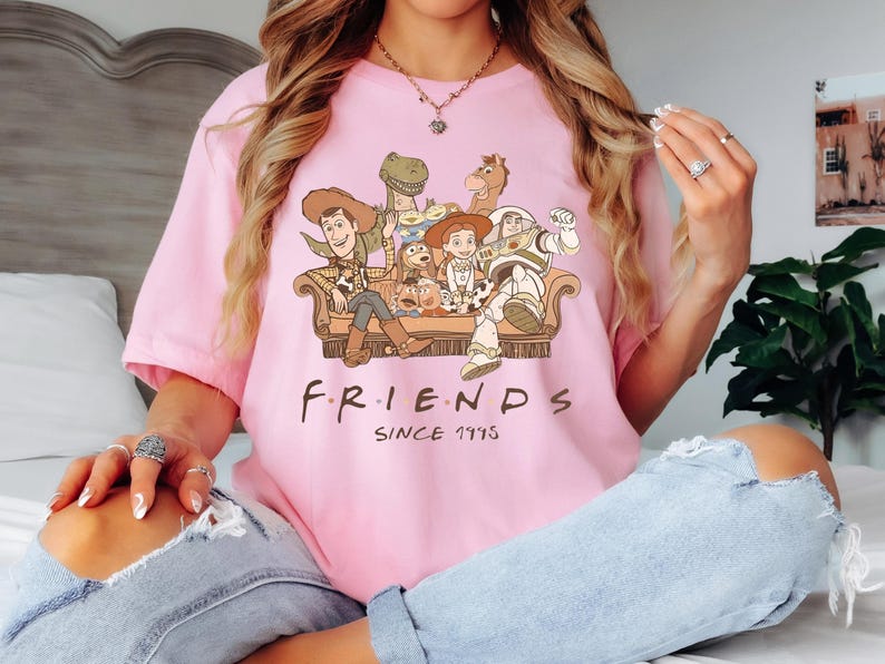 Op de afbeelding: Roze T-shirt met een afbeelding van de Toy Story-personages die op een bank zitten. De tekst "FRIENDS SINCE 1995" staat onder de afbeelding.