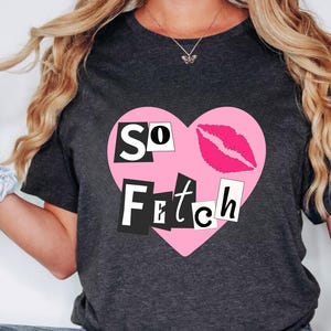 May include: A dark grey t-shirt with a pink heart graphic. The heart has the words "SO FETCH" in black letters and a pink lipstick print.