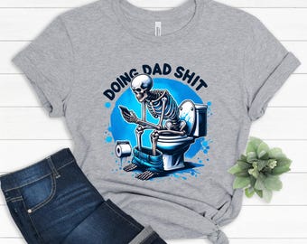 May include: A light gray t-shirt featuring a humorous graphic of a skeleton sitting on a toilet while using a smartphone. The text "DOING DAD SHIT" is displayed above the skeleton in a blue circle.