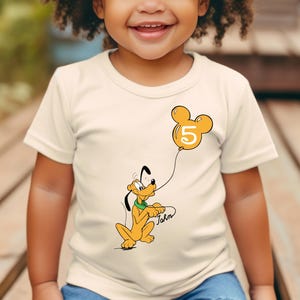 May include: A white t-shirt with a cartoon image of Pluto holding a yellow balloon with a Mickey Mouse ear shape and the number 5 on it. The text "John" is written below Pluto.