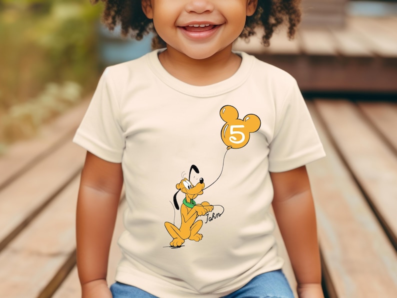 May include: A white t-shirt with a cartoon image of Pluto holding a yellow balloon with a Mickey Mouse ear shape and the number 5 on it. The text "John" is written below Pluto.