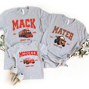 May include: Three gray t-shirts with Cars characters and text. The largest shirt says "MACK EST 1985 DISNEY CAR" with a red truck. The medium shirt says "MATER EST 1951 DISNEY CAR" with a brown tow truck. The smallest shirt says "MCQUEEN EST 1977 DISNEY CAR" with a red race car.